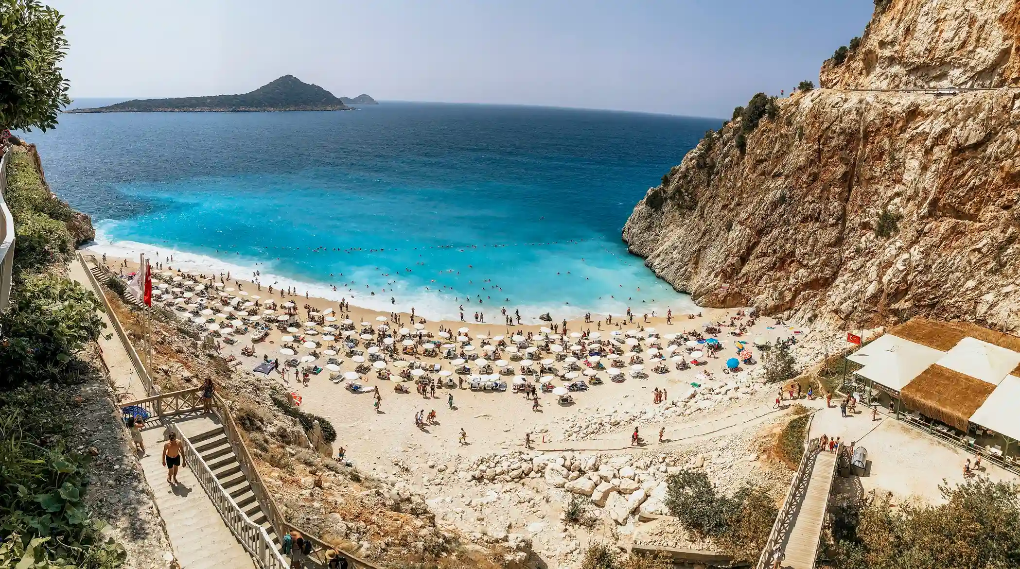 Turquoise beach at Antalya lined with sunbathers and cliffs, a Mediterranean gem recommended by icanfly for Turkey escapes.