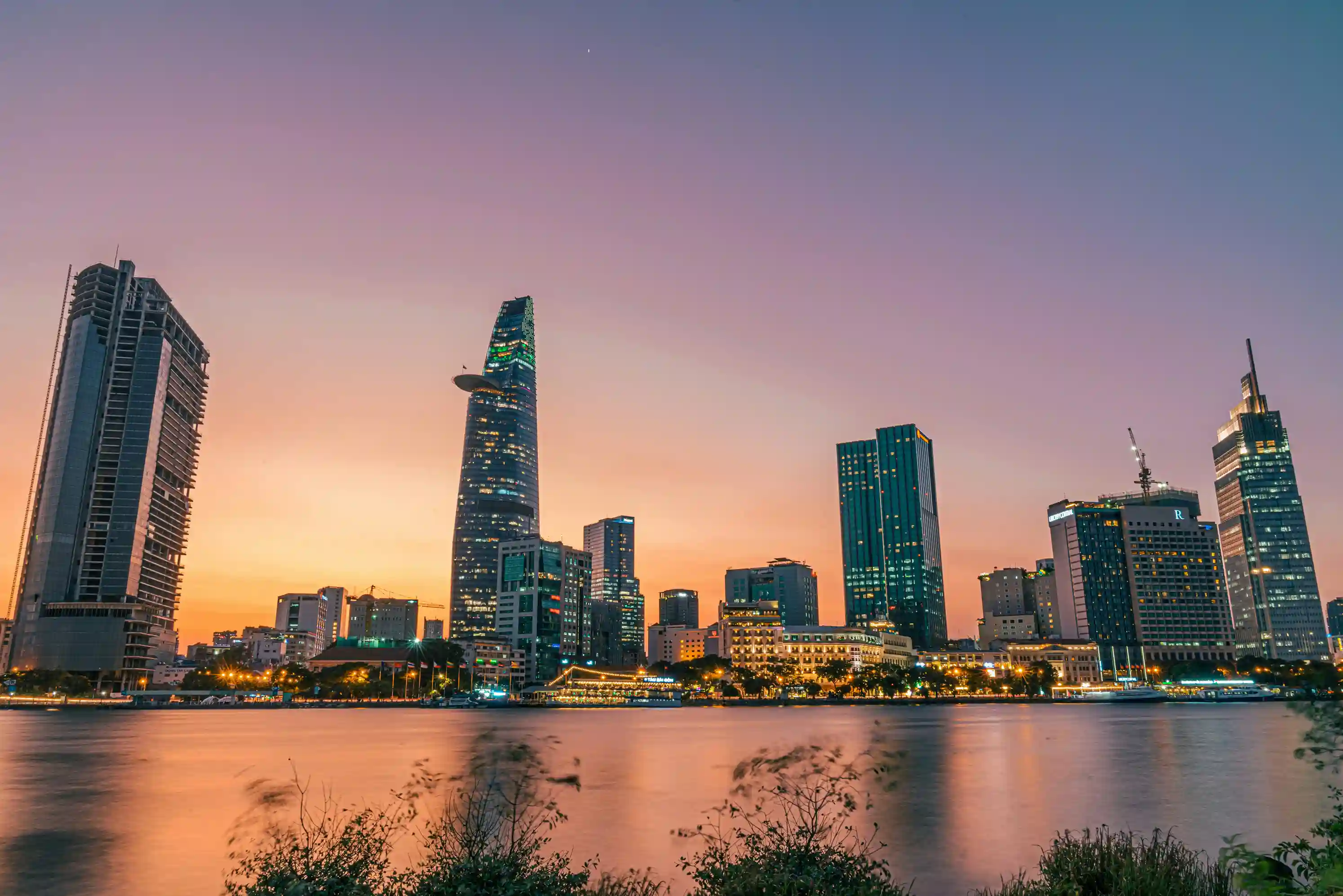 Vietnam city skyline at sunset with modern high-rises reflected on a calm river, showcasing a vibrant urban landscape promoted by icanfly as a top destination for your next escape.